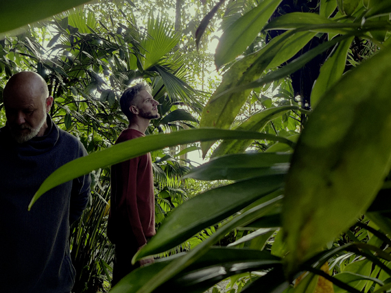 The Phyne boys, standing in the jungle, Sascha on the left-, Benj on the right side. Sascha looks to the ground, while Benj has his head turned right, looking to in to the distance (heroicly).